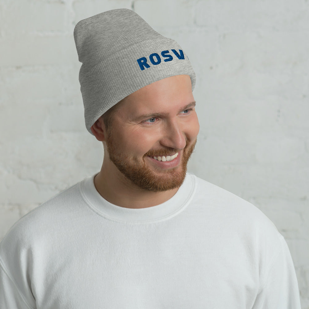 Hymyilevä partainen mies käyttää Meemirottan harmaata Rosvo Pipo -pipoa, jossa on sinisillä kirjaimilla "ROSV", joka on ihanteellinen ainutlaatuisia lahjaideavaihtoehtoja etsiville. Hänellä on myös valkoinen collegepaita ja hän seisoo valkoista tiiliseinää vasten.